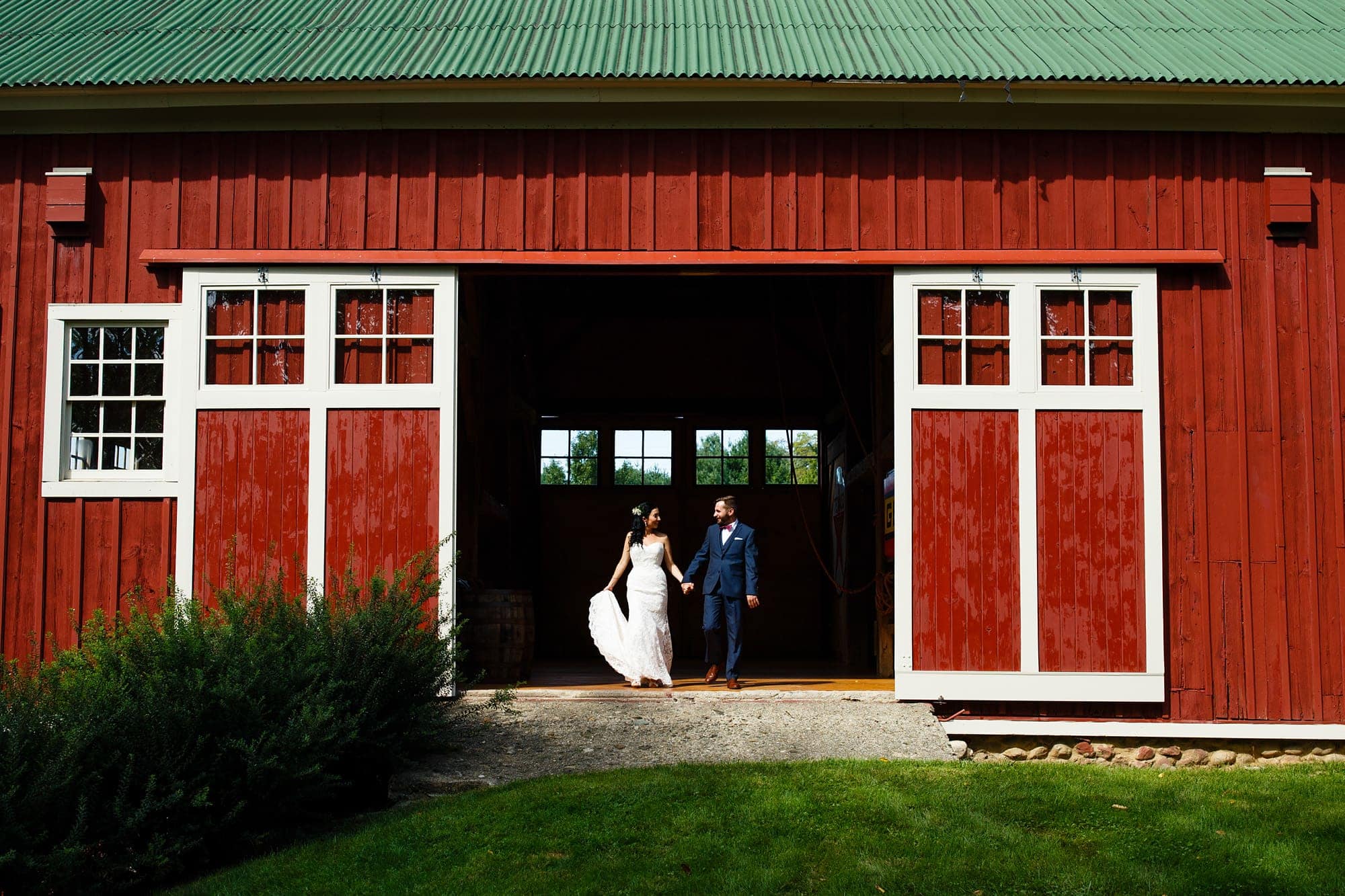 A couple walk hand in hand out of a red barn during a portrait session following their first look on a farm near Willow Harbor Vineyards in Three Oaks, Michigan.