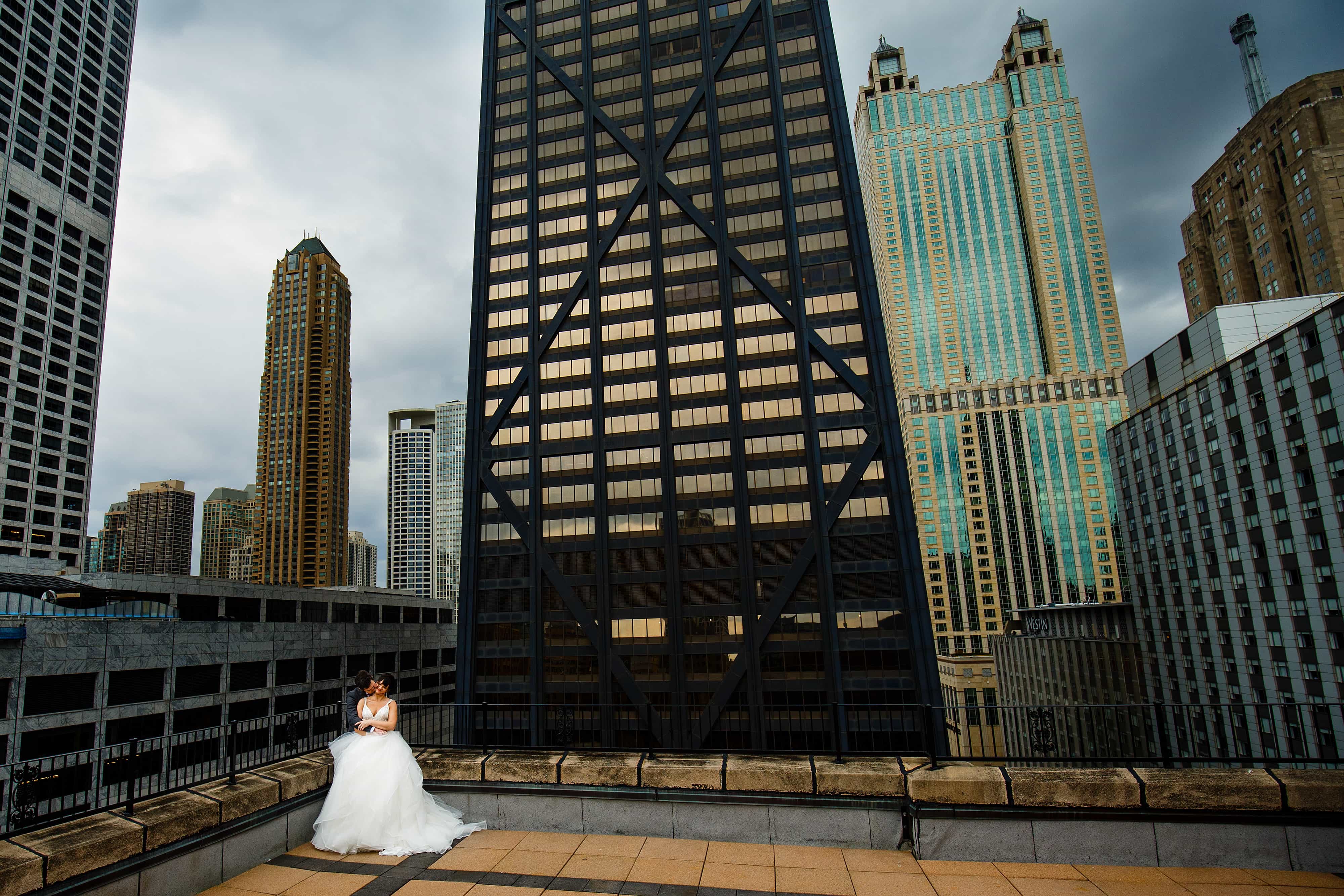 Chelsea and Ori embrace on a rooftop in downtown Chicago near the Hancock building