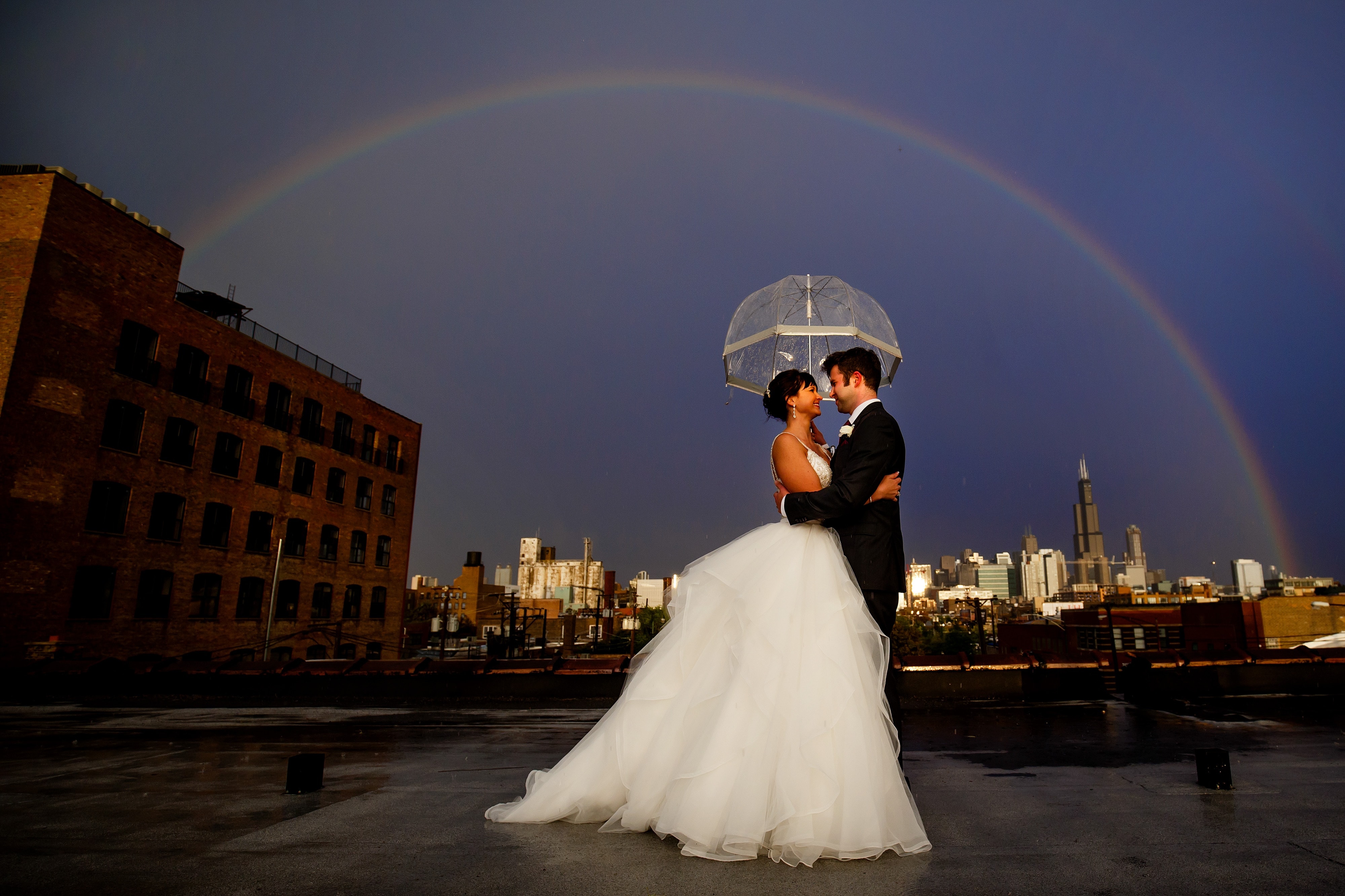Chelsea and Ori embrace under an umbrella as they're surrounded by a rainbow on the rooftop during their Room 1520 wedding in Chicago