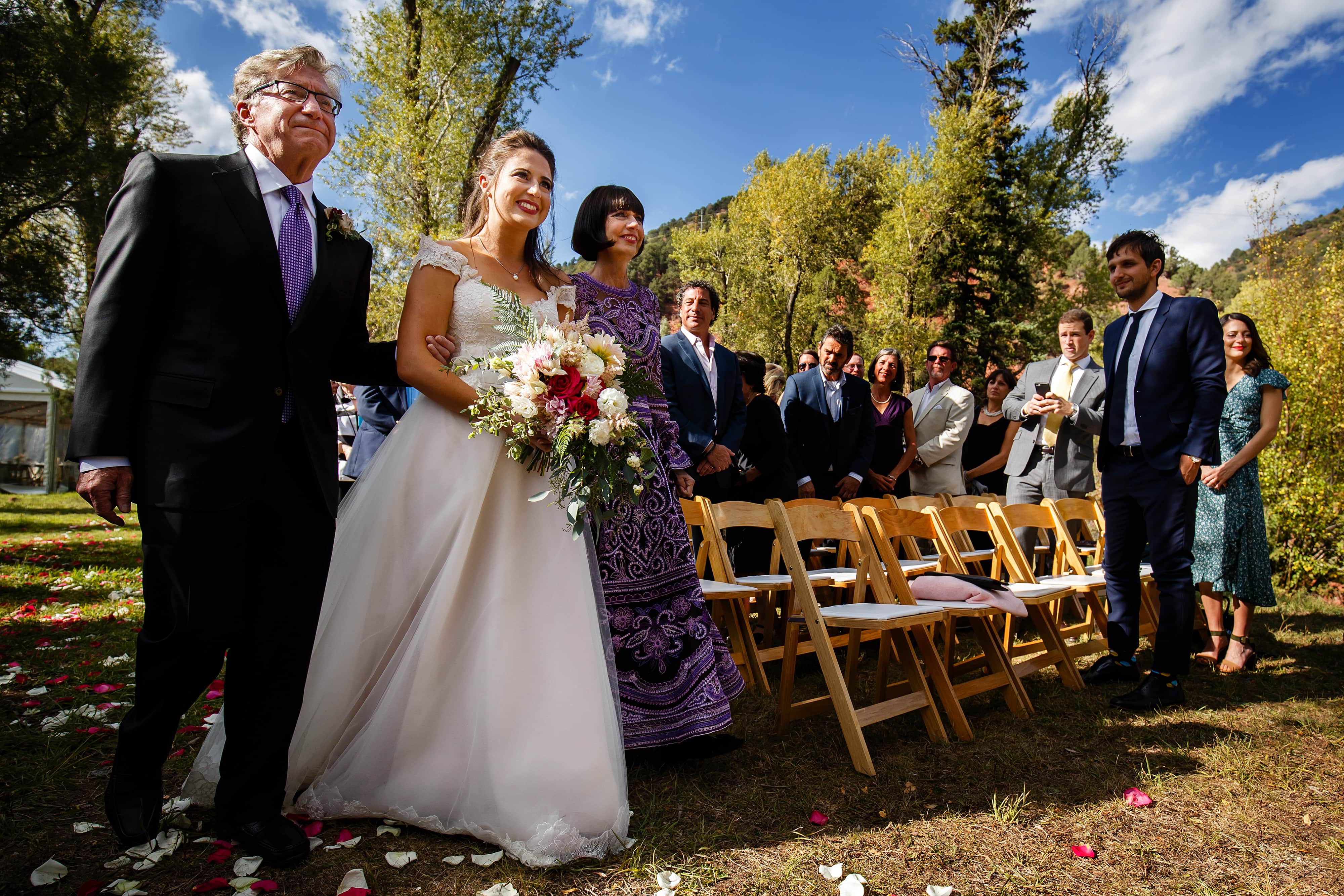 Mallory walks down the aisle as a stunning bride with her parents during the Snowmass Cottages wedding ceremony