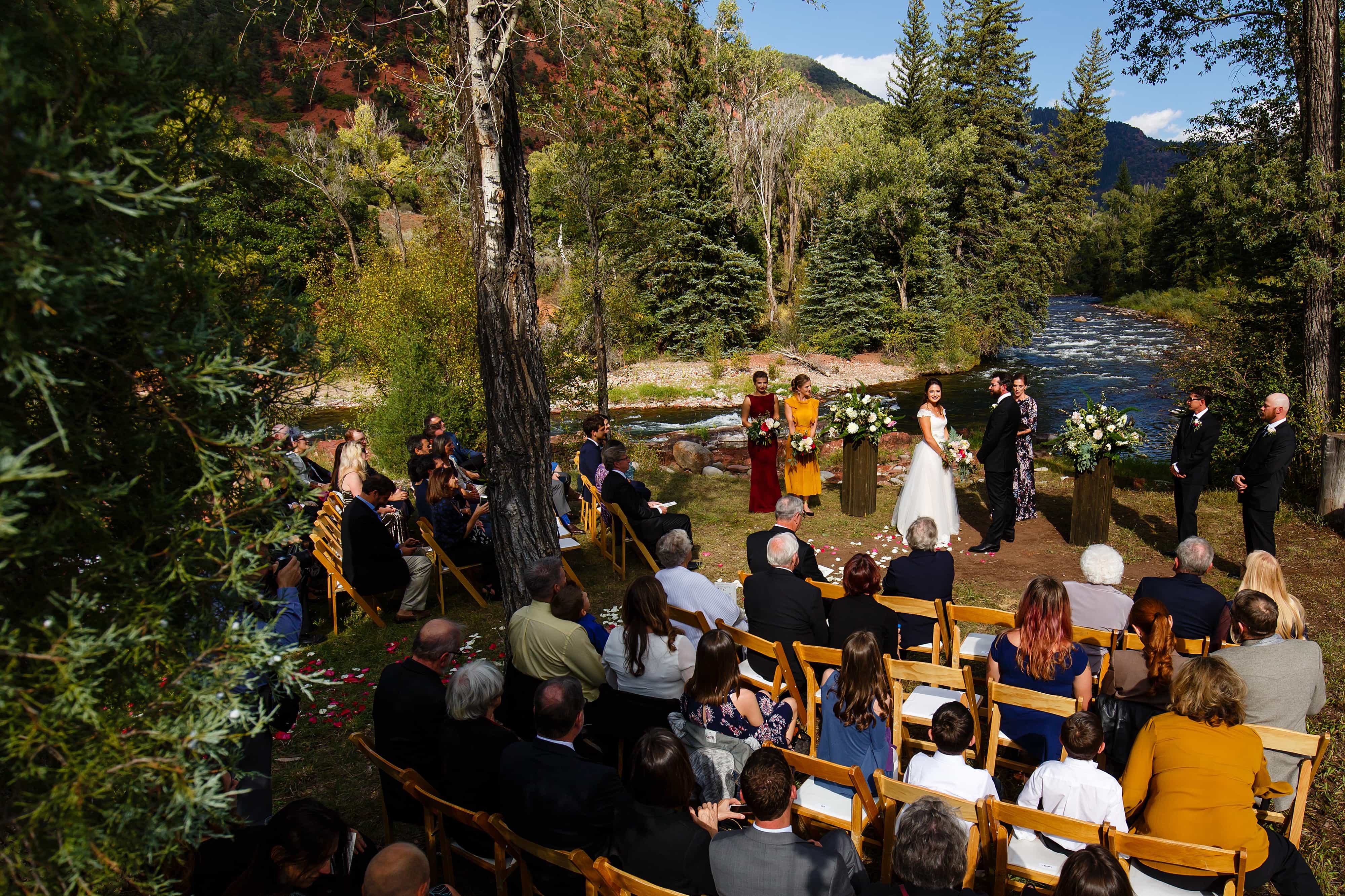The couple looks out to guests as the creek rushes in the background during their fall wedding ceremony at Snowmass Cottages in Colorado