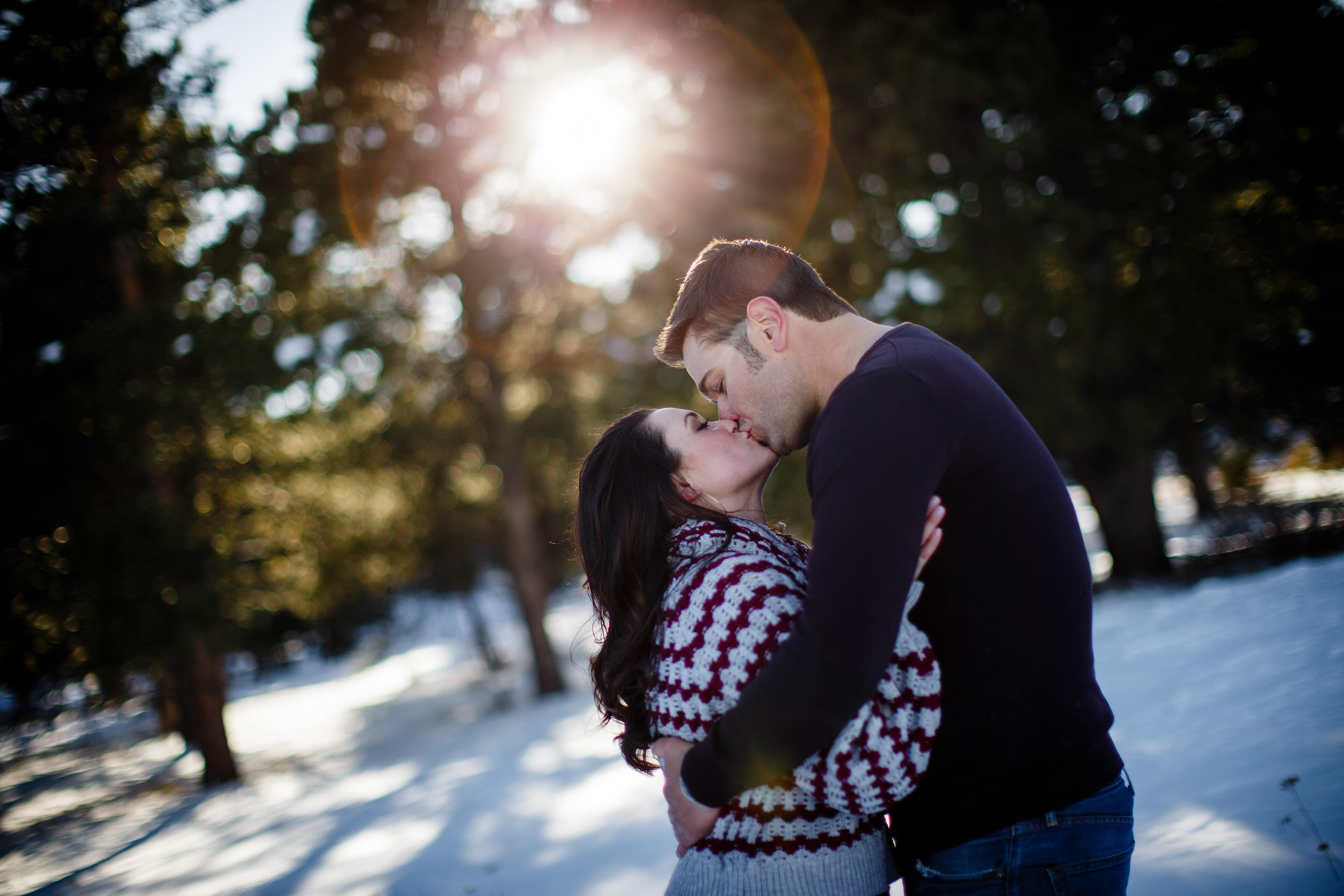 The sun flares through the trees as Jordan and Melissa share a kiss during their winter engagement session at Elk Meadow Park in Evergreen, Colorado
