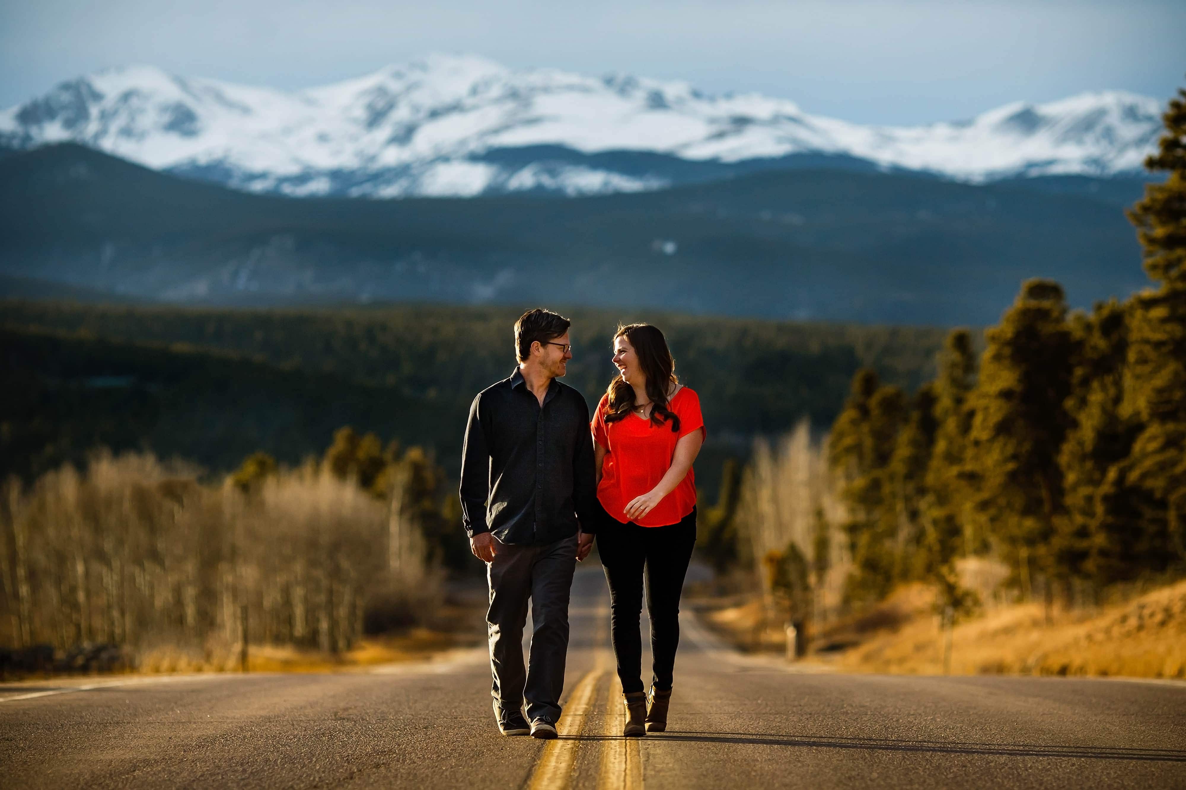 Brian and Kate hold hands and walk along Colorado highway 119 in Black Hawk, Colorado with the snow capped mountains seen in the background during their Spring Golden Gate Canyon State Park engagement