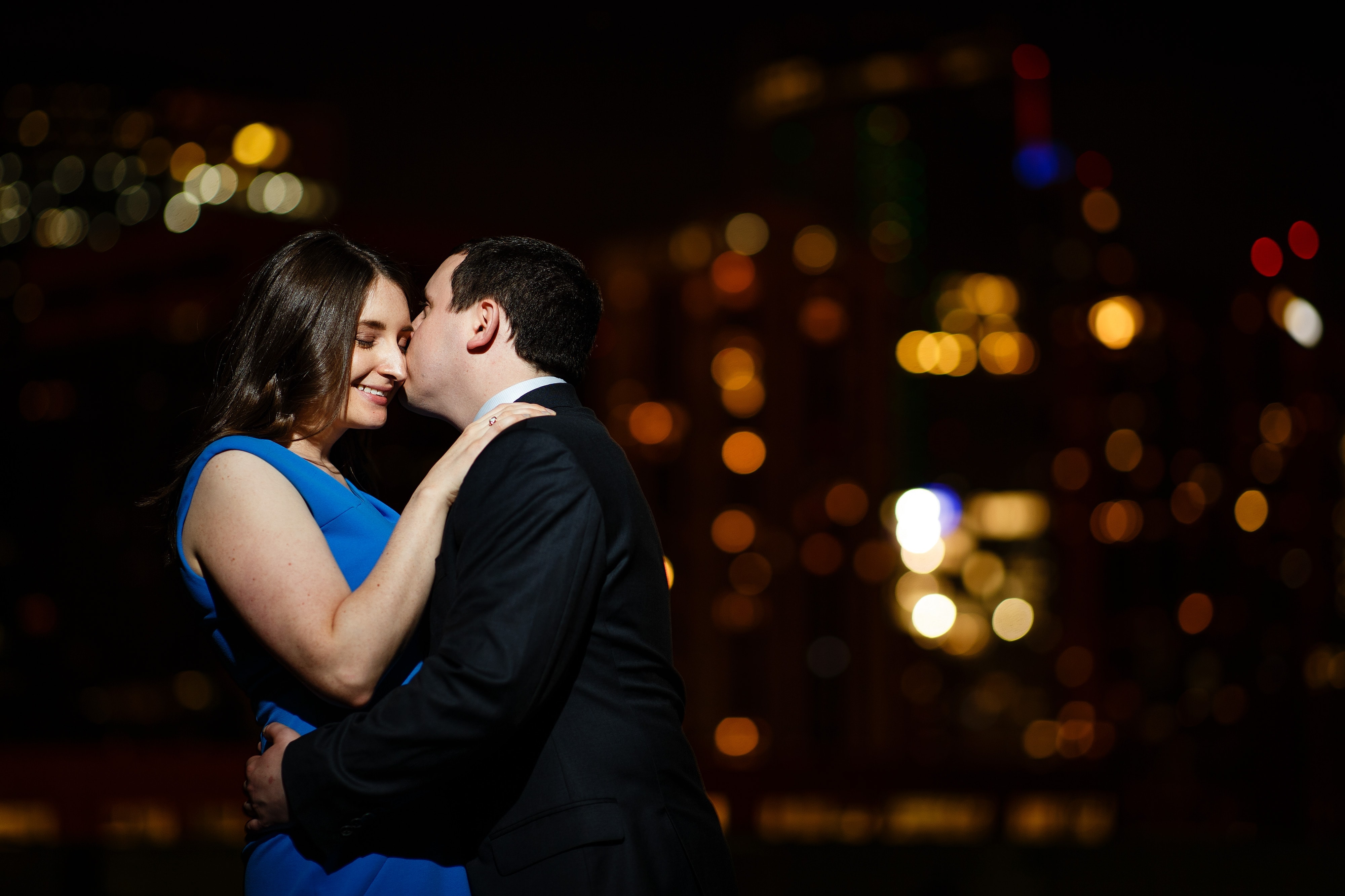 Joel kisses Katie with the Denver skyline in the background during their downtown Denver engagement session at night