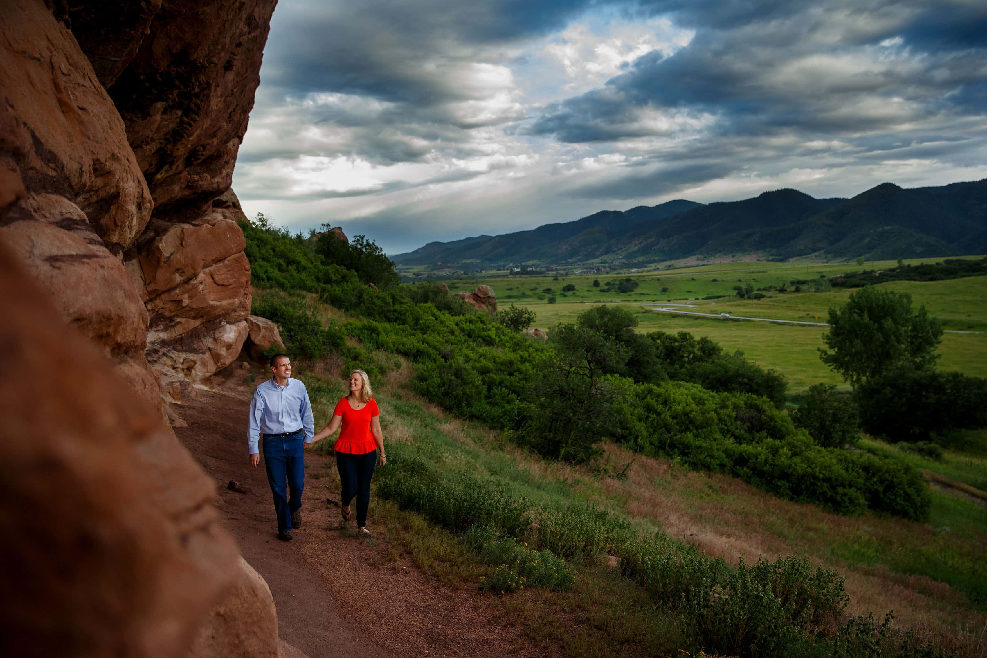 A couple walk together near a red rock formation at Roxborough State Park during their engagement session