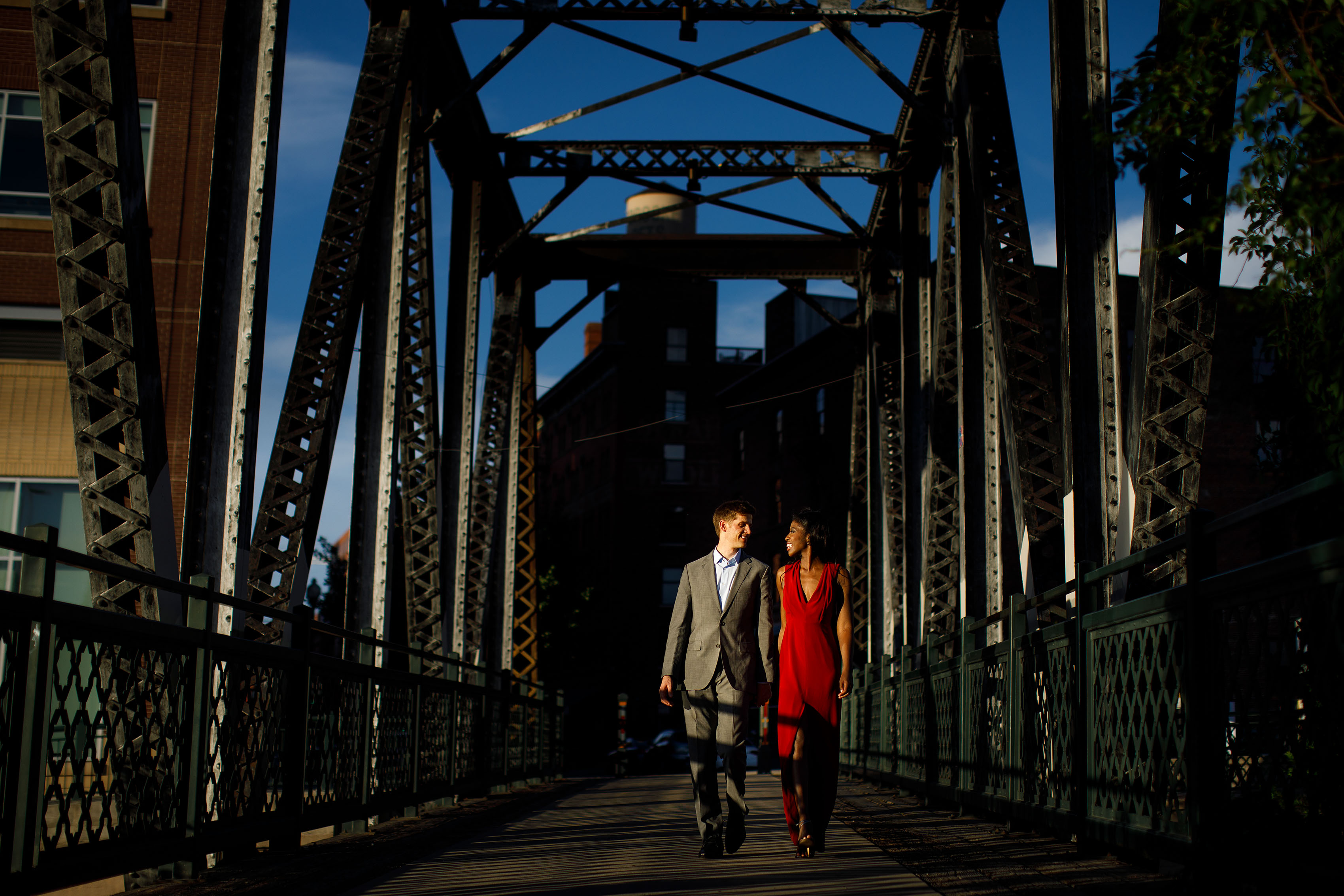 Man in a suit and woman in a red dress walk together at sunset on the Wynkoop Street Bridge