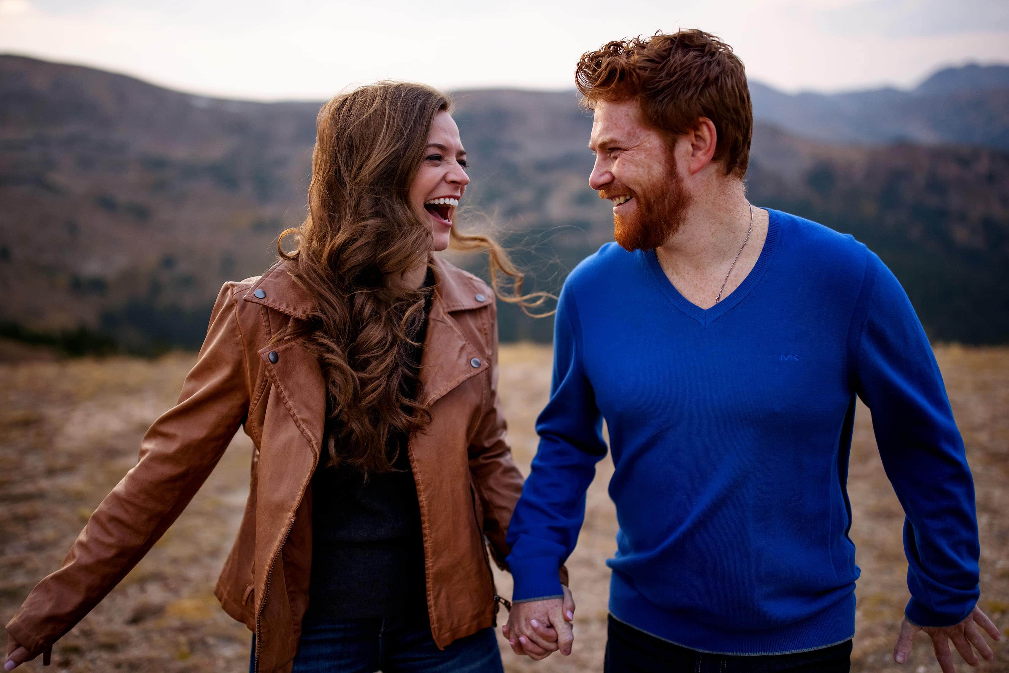 A couple laughs together on the top of a mountain during their engagement session